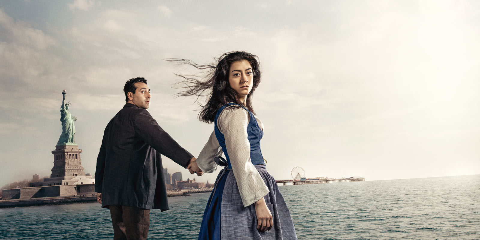 Kampanjbild till Utvandrarna, Gizem Erdogan och Ashkan Ghods håller varandra i handen i fonden ses frihetsgudinnan.